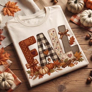 Op de afbeelding: Een crèmekleurig T-shirt met het woord FALL in grote letters. De F heeft een kabelgebreide textuur, de A is zwart-wit geblokt, de L is bloemig en de tweede L heeft een bruin pied-de-poule patroon. Versierd met pompoenen en bladeren.