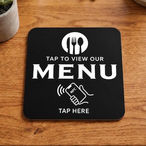 Pode incluir: Uma placa quadrada preta com gráficos e texto em branco. No topo, um círculo branco contém um ícone de faca, garfo e colher. Abaixo, o texto diz TAP TO VIEW OUR MENU. Um desenho de linha branca de uma mão segurando um telefone com o texto NFC aparece acima das palavras TAP HERE.