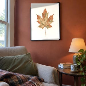 May include: A framed wall art print featuring a single maple leaf with a detailed texture in shades of orange, brown, and muted green against a plain white background. The artwork is enclosed in a thin black frame and displayed on a wall.
