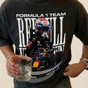 Pode incluir: Uma t-shirt de manga curta cinzento-carv&atilde;o com uma grande impress&atilde;o gr&aacute;fica de um carro de corrida de F&oacute;rmula 1 e um piloto com capacete. O texto diz FORMULA 1 TEAM, RED BULL, BORN TO RACE e TAPPEN, com pequenos detalhes impressos.