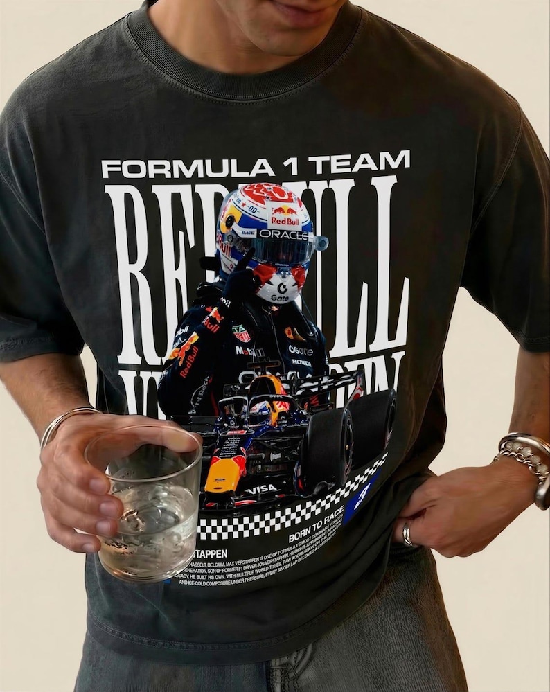 Pode incluir: Uma t-shirt de manga curta cinzento-carv&atilde;o com uma grande impress&atilde;o gr&aacute;fica de um carro de corrida de F&oacute;rmula 1 e um piloto com capacete. O texto diz FORMULA 1 TEAM, RED BULL, BORN TO RACE e TAPPEN, com pequenos detalhes impressos.