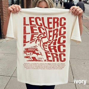 Pode incluir: Uma t-shirt de cor marfim com um gráfico grande, vermelho e distorcido do nome LECLERC repetido num padrão de grelha. Abaixo do texto encontra-se uma ilustração vermelha de um capacete de corrida de Fórmula 1 com o número 16 e texto impresso ilegível.