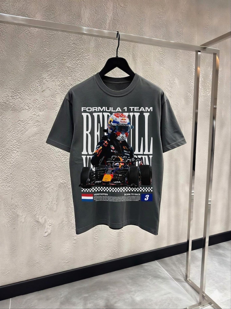 Pode incluir: Uma t-shirt cinzento-carv&atilde;o de manga curta com uma impress&atilde;o gr&aacute;fica de um carro de F&oacute;rmula 1 e um piloto. O texto diz FORMULA 1 TEAM, RED BULL, VERSTAPPEN, BORN TO RACE e o n&uacute;mero 3. Uma pequena bandeira holandesa est&aacute; impressa em baixo.