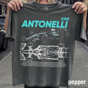 Pode incluir: Uma t-shirt de manga curta cinzento-carvão com um gráfico ciano do nome KIMI ANTONELLI sobre um carro de corrida de Fórmula 1. Abaixo do carro, há um desenho técnico branco do veículo e um parágrafo de texto sobre a carreira do piloto.