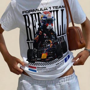 Pode incluir: Uma t-shirt branca de manga curta com uma grande impress&atilde;o gr&aacute;fica de um carro de F&oacute;rmula 1 e um piloto com capacete. O texto inclui "FORMULA 1 TEAM", "RED BULL", "VERSTAPPEN" e "BORN TO RACE" com um padr&atilde;o de bandeira axadrezada.