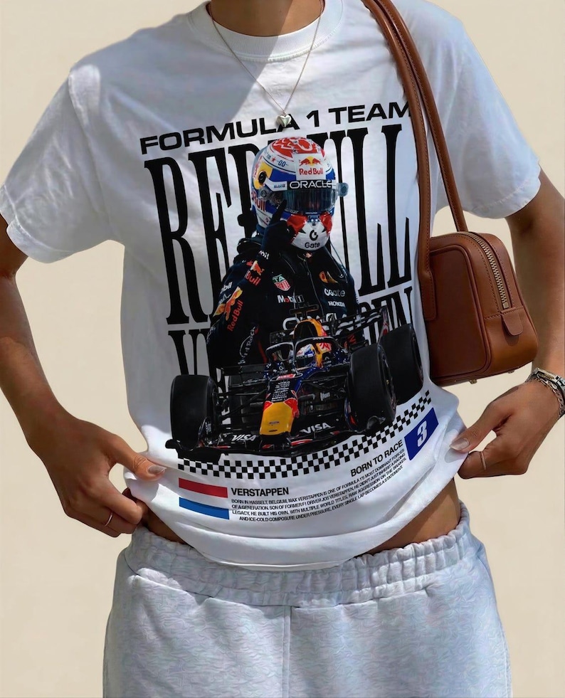 Pode incluir: Uma t-shirt branca de manga curta com uma grande impress&atilde;o gr&aacute;fica de um carro de F&oacute;rmula 1 e um piloto com capacete. O texto inclui "FORMULA 1 TEAM", "RED BULL", "VERSTAPPEN" e "BORN TO RACE" com um padr&atilde;o de bandeira axadrezada.