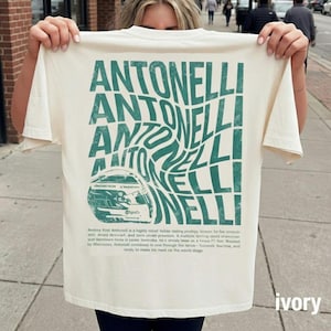 以下が含まれることがあります： アイボリー色のTシャツで、背面に大きなダークグリーンのグラフィックがプリントされています。歪んだ波状のフォントで繰り返される「ANTONELLI」という文字と、レーシングヘルメットの線画、そしてその下に小さなテキストがデザインされています。