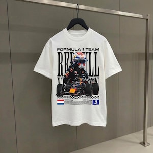 Pode incluir: Uma t-shirt branca de manga curta com uma impress&atilde;o gr&aacute;fica de um carro de corrida de F&oacute;rmula 1 e um piloto. O texto diz Formula 1 Team, Red Bull, Verstappen, Born to Race e o n&uacute;mero 3, acompanhado por uma bandeira holandesa e um padr&atilde;o xadrez.