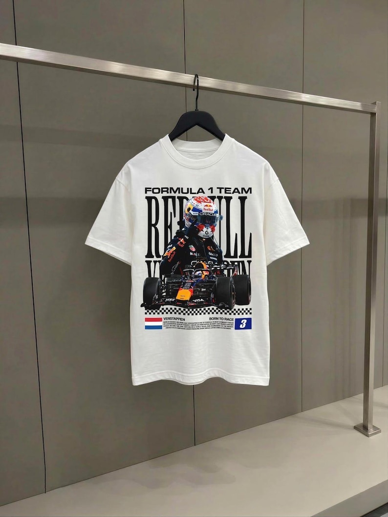 Pode incluir: Uma t-shirt branca de manga curta com uma impress&atilde;o gr&aacute;fica de um carro de corrida de F&oacute;rmula 1 e um piloto. O texto diz Formula 1 Team, Red Bull, Verstappen, Born to Race e o n&uacute;mero 3, acompanhado por uma bandeira holandesa e um padr&atilde;o xadrez.