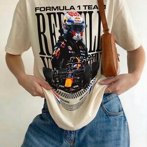 Pode incluir: Uma t-shirt de manga curta cor de creme com uma grande impress&atilde;o gr&aacute;fica de um carro de F&oacute;rmula 1 e um piloto com capacete. O texto diz FORMULA 1 TEAM, RED BULL e VERSTAPPEN, acompanhado por um padr&atilde;o de xadrez e cal&ccedil;as de ganga azuis.
