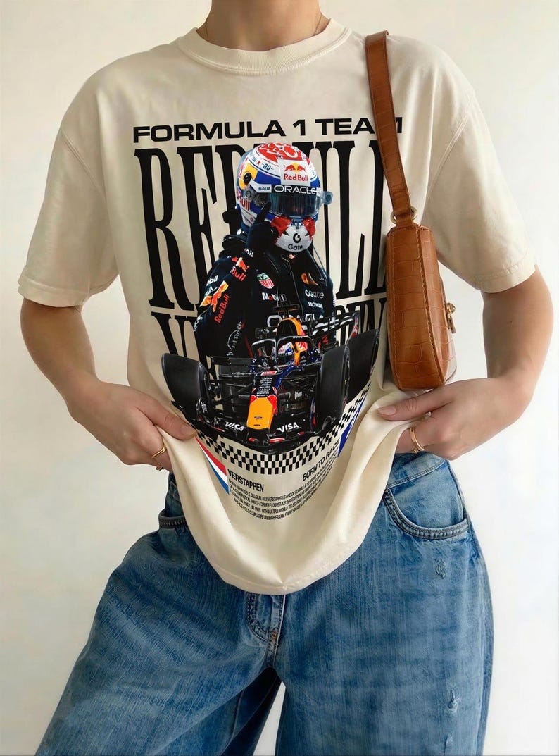 Pode incluir: Uma t-shirt de manga curta cor de creme com uma grande impress&atilde;o gr&aacute;fica de um carro de F&oacute;rmula 1 e um piloto com capacete. O texto diz FORMULA 1 TEAM, RED BULL e VERSTAPPEN, acompanhado por um padr&atilde;o de xadrez e cal&ccedil;as de ganga azuis.
