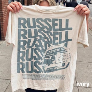 Pode incluir: Uma t-shirt de manga curta na cor marfim com a palavra RUSSELL repetida em letras grandes azul-escuras. Um gráfico de um capacete de corrida com o número 63 e texto incluindo SOLERA, CROWDSTRIKE e AKKODIS está impresso abaixo.