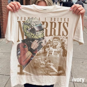 Pode incluir: Uma t-shirt cor de marfim com uma estampa gráfica do piloto de Fórmula 1 Lando Norris. O design inclui um capacete grande, um retrato do piloto e um carro de corrida. O texto diz FORMULA 1 DRIVER NORRIS e o número 1 está impresso abaixo.