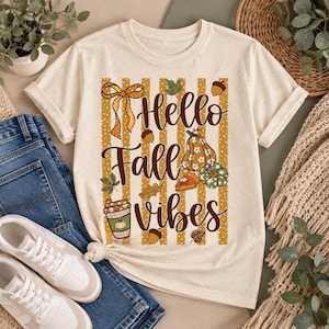 Pode incluir: Uma t-shirt cor de creme com uma estampa gráfica de listras verticais amarelo-mostarda e bolinhas brancas. O design inclui o texto "Hello Fall Vibes" em letra cursiva castanha, rodeado por ilustrações de um laço, folhas, bolotas, uma abóbora e uma chávena de café.