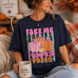 Puede incluir: Una camiseta azul marino con un estampado gráfico de tres calabazas en rosa y naranja con patrones de lunares y cuadros. El texto "TAKE ME to the PUMPKIN PATCH" aparece en letras rosas y naranjas sobre un fondo de rayas verde azulado con pequeñas estrellas doradas.