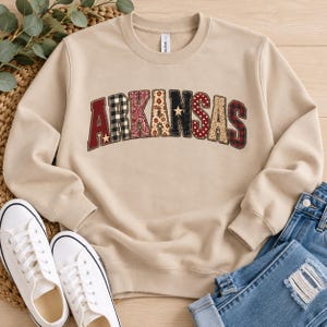 May include: A beige crewneck sweatshirt featuring the word ARKANSAS in an arched, multi-patterned applique font. Each letter displays a different texture, including red, black and white plaid, leopard print, and polka dots with small star accents.