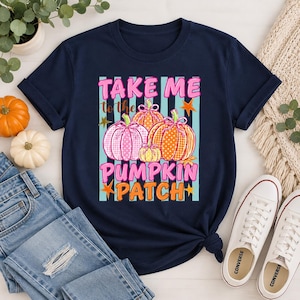 Könnte beinhalten: Ein dunkelblaues Kurzarm-T-Shirt mit einem Grafikdruck von drei gemusterten Kürbissen in Pink und Orange. Der Text "TAKE ME to the PUMPKIN PATCH" ist in pinken und orangen Buchstaben vor einem Hintergrund mit hellblauen vertikalen Streifen gedruckt.