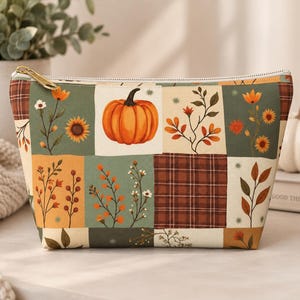 May include: A zippered pouch featuring a patchwork design with autumn-themed illustrations including an orange pumpkin, brown plaid squares, and various floral and botanical motifs in shades of green, orange, yellow, and cream on a fabric surface.