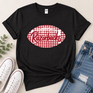 May include: A black short-sleeved t-shirt featuring a central graphic of a football shape with a red and white gingham pattern. The word Razorbacks is printed in a red script font across the center of the football graphic on the shirt.