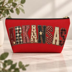 May include: A red zippered pouch featuring the word ARKANSAS in large, fabric-appliqued letters. Each letter displays a unique pattern, including black and white buffalo plaid, red floral, cream polka dots, dark denim, and red plaid, with gold stars.