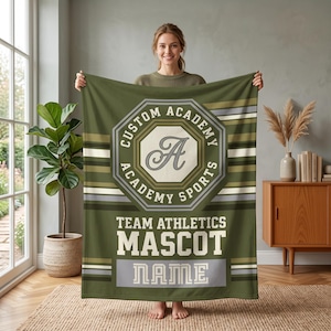 May include: An olive green fleece blanket featuring a central octagonal emblem with the text CUSTOM ACADEMY ACADEMY SPORTS and a large letter A. Below the emblem, the text reads TEAM ATHLETICS MASCOT and NAME on a light grey rectangular background.