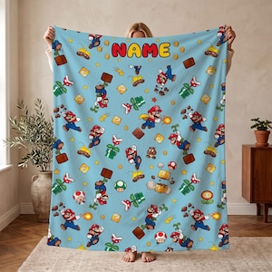 Könnte beinhalten: Eine hellblaue Fleecedecke mit einem wiederkehrenden Muster aus Mario-Figuren, Piranha-Pflanzen, Pilzen und Goldmünzen. Das Wort NAME ist in großen, rot gefüllten Buchstaben mit gelber Umrandung oben in der Mitte aufgedruckt.