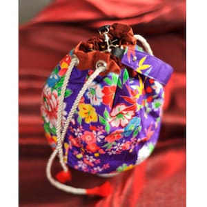 Può includere: Una borsa a secchiello con coulisse viola caratterizzata da un vivace motivo floreale con fiori rossi, rosa, gialli e bianchi. La borsa include una fodera interna in tessuto marrone, un manico viola e spesse corde bianche con nappe rosse.