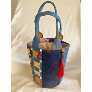 Op de afbeelding: Een emmertas gemaakt van blauwe spijkerstof met twee handvatten. De buitenkant heeft een patchwork-ontwerp met diverse kleurrijke stoffen vierkantjes en een felrode kwast aan de zijkant. De binnenzijde is gevoerd met een lichte stof.