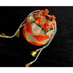 Op de afbeelding: Een buidel met trekkoord van levendige oranje en gouden metallic stof met een geometrisch patroon. De tas is aan de bovenkant dichtgetrokken met witte gevlochten koorden die eindigen in kleine, glanzende gouden kwastjes voor een decoratieve sluiting.