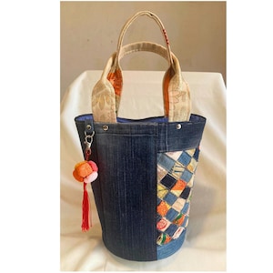 May include: A bucket-style handbag constructed from dark blue denim fabric, featuring a side panel of multicolored patchwork squares in various textures. The bag has two beige patterned handles and a hanging charm with a red tassel and fabric flower.