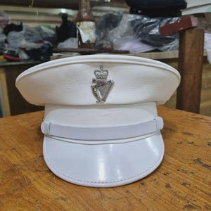 May include: A white peaked cap featuring a silver-toned metal emblem on the front band. The emblem depicts a crown positioned above a harp. The cap has a structured crown, a horizontal band with a chin strap, and a smooth, glossy white visor.