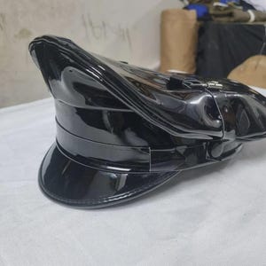 May include: A black patent leather cap with a high-gloss finish, featuring a structured crown, a wide horizontal band, and a curved visor. The cap includes a decorative strap across the front secured by small circular buttons on each side.