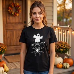 Pode incluir: Uma t-shirt preta de manga curta com uma impressão gráfica branca de um fantasma sobre uma teia de aranha com uma aranha no centro. O texto THE BOO CREW está impresso em letras maiúsculas brancas entre o fantasma e a teia.