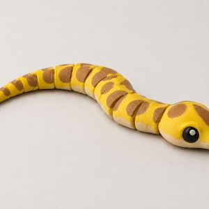 Könnte beinhalten: Eine gelb-braune, bewegliche Schlangenfigur mit segmentiertem Körper. Die Schlange hat eine leuchtend gelbe Grundfarbe mit abgerundeten braunen Flecken auf dem Rücken und große, dunkle, glänzende Augen auf ihrem abgerundeten Kopf.