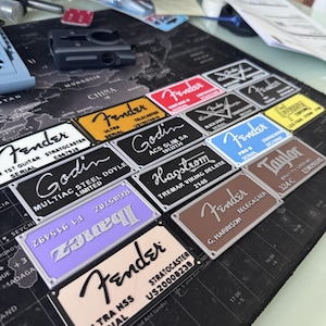 May include: A collection of rectangular metal guitar neck plates in various colors including white, yellow, red, black, blue, brown, and purple. Each plate features engraved brand names like Fender, Godin, Hagstrom, Taylor, and Ibanez with serial numbers.