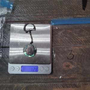 May include: A circular keychain featuring an iridescent abalone shell inlay with a central metallic profile relief, attached to a dark metal chain and ring, resting on a digital scale displaying a weight of 12.8 grams.