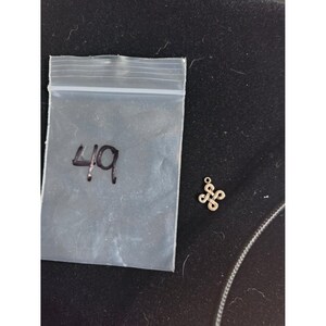May include: A small, metallic, four-looped knot charm with a jump ring at the top, positioned next to a clear plastic baggie marked with the handwritten number 49 in black ink. The charm features a symmetrical, interwoven design.
