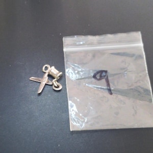 May include: A silver-toned metal charm shaped like a pair of scissors with a small loop at the top for attachment. The charm is positioned next to a small, clear plastic resealable bag that has a dark handwritten mark on its surface.