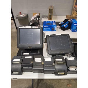 May include: Two black point-of-sale touch screen monitors positioned side-by-side, accompanied by six black thermal receipt printers arranged in a row. The equipment is set on a white table, with various boxes and a blue tool bag visible in the background.