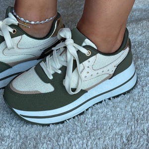 Può includere: Un paio di sneakers color verde oliva e crema con una spessa suola platform bianca, pannelli in mesh e sintetici, lacci bianchi e dettagli color oro metallizzato sul tallone e sugli occhielli. Il design include piccole borchie decorative laterali.