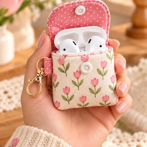 May include: A fabric protective case for wireless earbuds featuring a cream background with a repeating pink tulip pattern and green stems. The case has a pink and white polka dot interior lining, a white snap button closure, and a gold metal keychain.