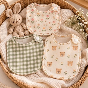 Pode incluir: Três babadores de bebê com bordas arredondadas e fechos de pressão brancos. Um babador apresenta um padrão floral rosa e laranja em fundo creme, um tem xadrez verde e branco, e o terceiro exibe um padrão de pequenos ursos castanhos e ramos.