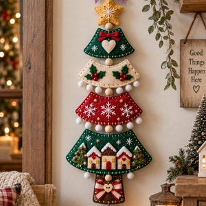 Puede incluir: Un tapiz vertical de fieltro con forma de árbol de Navidad, con segmentos apilados en verde, crema y rojo. Está decorado con un lazo morado, una estrella amarilla, pompones blancos, copos de nieve bordados, acebo y una pequeña escena de pueblo.