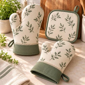 May include: Two oven mitts and one square pot holder featuring an off-white fabric with a green leaf pattern. Each piece has a solid green cuff and a decorative cream-colored lace trim separating the patterned section from the cuff.