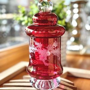 May include: A ruby red glass jar with a clear glass lid featuring a spherical finial. The cylindrical body is decorated with an etched white floral pattern and a row of small dots around the shoulder, resting on a clear, thick glass base.
