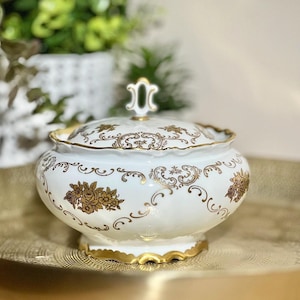 Może przedstawiać: Biała porcelanowa cukiernica z pokrywką, ozdobiona złotymi wzorami kwiatowymi i misternymi zawijasami. Cukiernica posiada falowaną podstawę i krawędź, obie wykończone złotym zdobieniem. Na środku pokrywki znajduje się mały, dekoracyjny uchwyt.