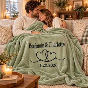Puede incluir: Una manta de felpa color verde salvia con el texto bordado en negro Benjamin & Charlotte, un gráfico de dos corazones entrelazados y la fecha 20.11.2026 centrados en la superficie de la tela.