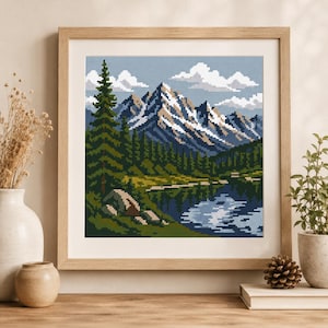 May include: A square cross-stitch embroidery depicting a mountain landscape with snow-capped peaks, a dense forest of green pine trees, and a reflective blue lake in the foreground, all contained within a light-coloured wooden frame.