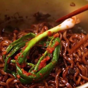 May include: A bowl of dark brown, wavy noodles coated in a thick sauce, topped with a single whole green onion stalk coated in a vibrant red chili paste, with a pair of wooden chopsticks visible lifting the onion from the noodle mixture.