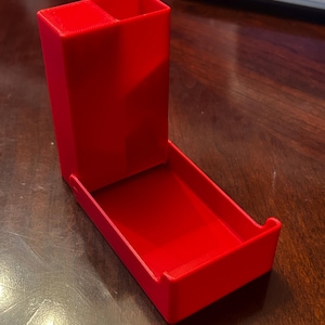May include: A bright red, 3D-printed plastic dice tower featuring a tall, vertical chute with a divided top opening and an attached horizontal tray at the base. The structure has a smooth, matte finish and a simple, functional geometric design.